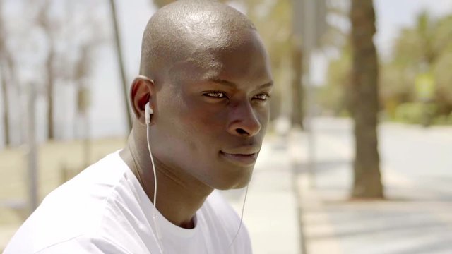 Handsome young African man listening to music on a set of earplugs as he sits outdoors an a seafront promenade