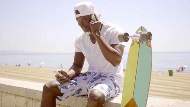 Black man checking his phone while seated on wall besides skateboard and wearing shorts and headset around his neck near the ocean and row of trees.