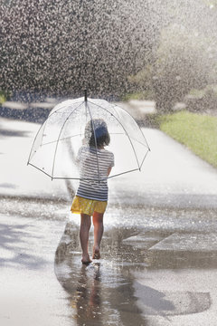 Rear view of barefoot girl carrying umbrella walking through street puddle