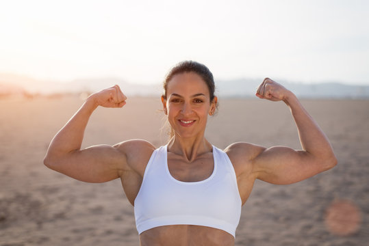 Young fitness woman flexing big strong biceps muscles towards the sun at urban beach. Cheerful female bodybuilder showing arms. Workout success concept.