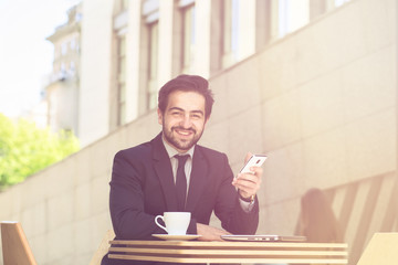Toned picture of businessman smiling while sitting in restaurant or cafe and working. Freelance man in black suit using mobile or smart phone.