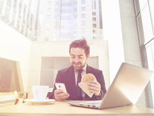 Toned picture of handsome businessman in black business suit using mobile phone while eating hamburger. Laptop computer is in front of him.