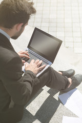Toned image of businessman using laptop computer while sitting on stairs in city centre. Blank screen on computer for any purposes.