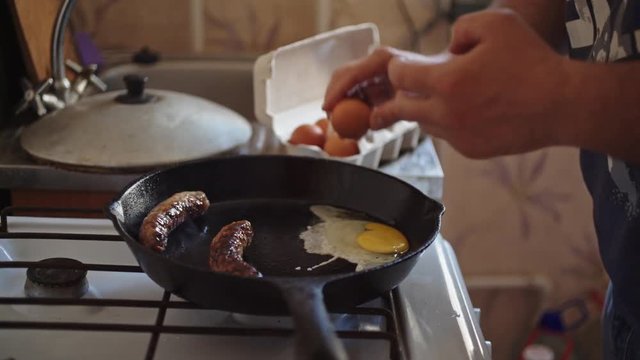 Man cooks breakfast of 4 eggs and 2 sausages in a large pan in the kitchen
