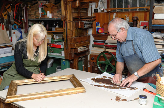 Senior man and young woman restoring picture frame and book spine in traditional bookbinding workshop