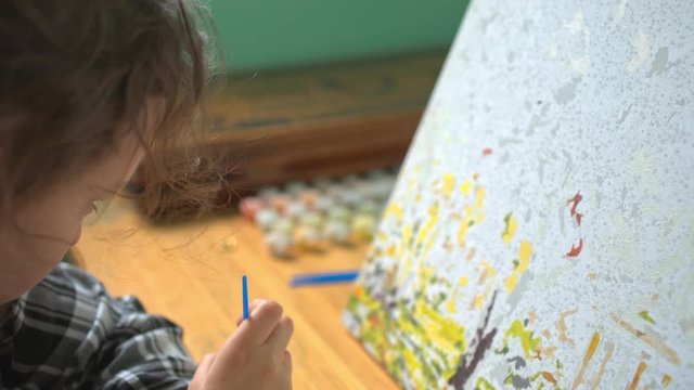 8-year-old girl in plaid shirt enthusiastically draws something on a large canvas by the window and dips a brush into the jar of paint. Close-up shots