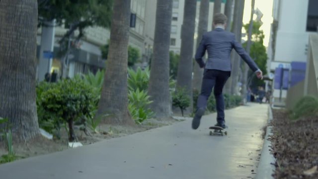 Young man in business suit rides away from the camera on a skateboard against a city background with traffic and trees in Downtown Los Angeles. Real time recorded at 24fps.