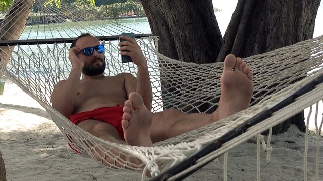 Young man using smartphone lying on hammock on beach
