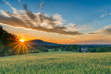 A perfect Spring day ends as the sun sets and sprays warm golden light over a beautiful farm with fields sprouting, a stone house, and mountain ridges in the background. - obrazy, fototapety, plakaty
