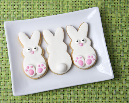 Three Home made designer Easter Bunny Cookies on rectangular plate with green placemat, two baby bunnies facing forward and one facing away with tail