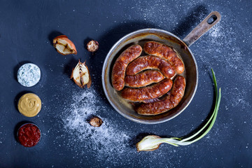 Tasty oven-baked sausages in a frying pan, three meat sauce, and grilled onion on a dark background. 