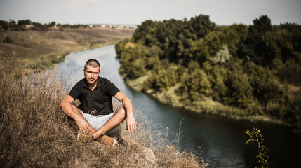 a bearded man in a black baseball cap, black shirt and shorts playing with white fluffy cat on top of a cliff on the background of the river and the forest