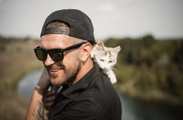 a bearded man in a black baseball cap, black shirt and shorts playing with white fluffy cat on top of a cliff on the background of the river and the forest