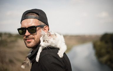 a bearded man in a black baseball cap, black shirt and shorts playing with white fluffy cat on top of a cliff on the background of the river and the forest