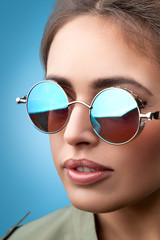 Close-up face portrait of young beautiful woman with perfect skin in round sunglasses smiling and looking at camera on the blue background. Beauty face make-up. 