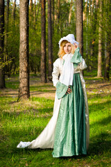  Fashionable shooting of a young short haired blond woman posing in the forest park wearing fancy empire style dress holding flowers. Concept of fantasy and magic.