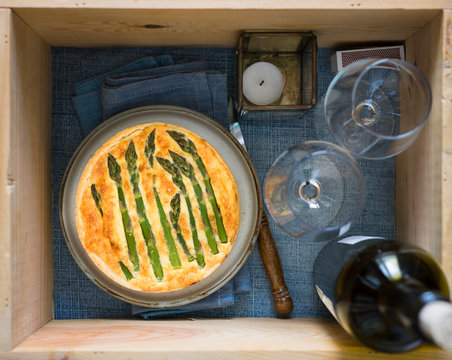 Wooden box prepared for picnic: asparagus tart, two wine glasses, wine bottle, candle. Selective focus on tart.