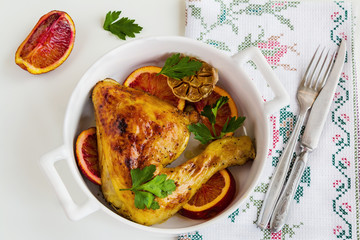 Roasted chicken legs, red oranges, garlic and parsley in white baking dish on white table. Cooked with sauce from orange, honey and olive oil. Top view.
