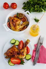 Roasted chicken legs cooked with sauce from orange, honey and olive oil. On white plate with fresh vegetables. White table. Top view.