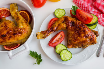 Roasted chicken legs cooked with sauce from orange, honey and olive oil. On white plate with fresh vegetables. White table. Top view.