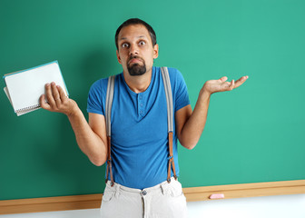 Doubting teacher spreads his hands looking at camera. Photo of adult man weighing decision near blackboard. Education concept