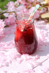 A jar of fresh raspberry jam on light rustic table covered with cherry flowers, romantic breakfast background, close up, in the garden