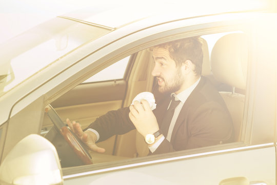 Toned picture of young businessman eating junk food while driving to work to office. Handsome man in black business suit looking to traffic jam.