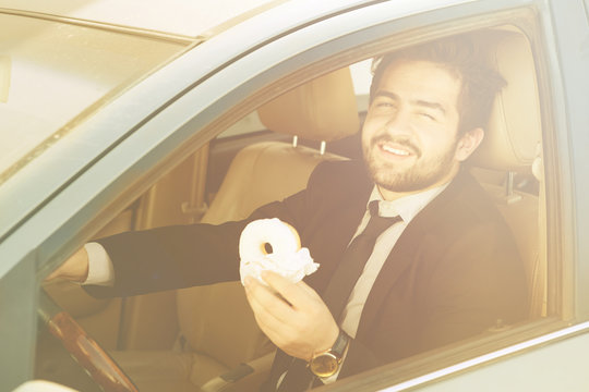 Toned picture of happy businessman eating doughnut and driving car to his office. Freelance man in black business suit smiling.