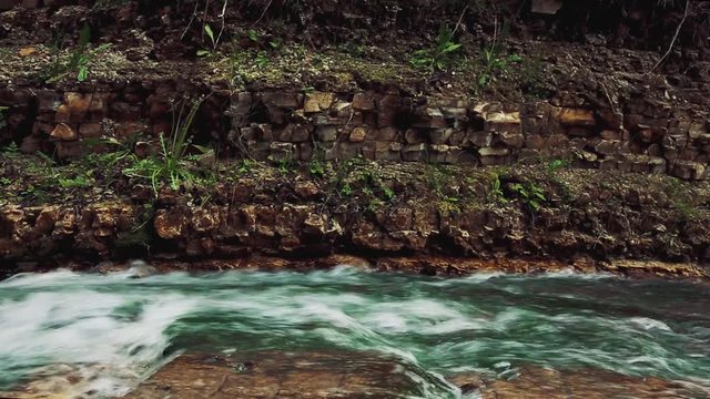 Wide shot of a yellow bird hopping along the top of a rock dam with a pool and waterfall