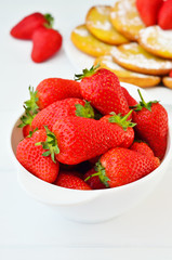 Full white bowl of strawberries on white table. Pancakes in the background