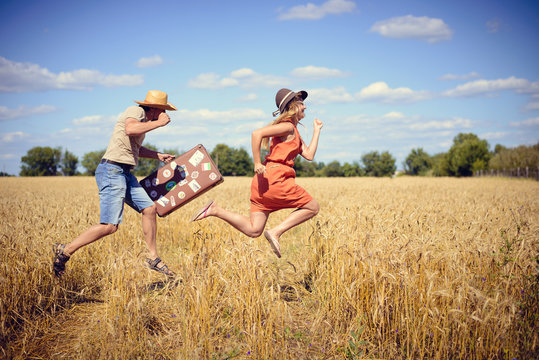 Joyful young couple having fun in wheat field. Excited man and woman running with retro leather suitcase on blue sky outdoor