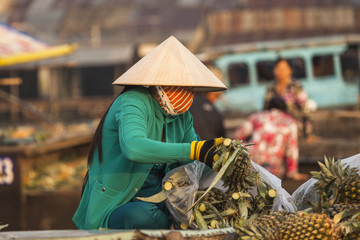 Vietnamese woman with typical conical hat ( sugegasa),  cut pineapple fruits in the  Cai Rang floating market on the Mekong delta. Can Tho, Vietnam - obrazy, fototapety, plakaty