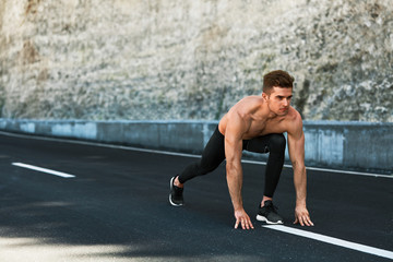 Fototapeta na wymiar Sports. Healthy Athletic Man With Fit Muscular Body In Starting Position For Running On Road. Handsome Runner Ready To Start Sprint Race. Fitness Model Training Outdoors In Summer. Workout Concept