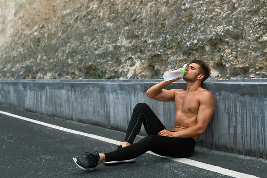 Tired Exhausted Athletic Man With Muscular Body Drinking Water, Resting After Running Workout. Thirsty Male Drinking Refreshing Drink After Outdoor Training On Hot Summer Day. Sports, Fitness Concept