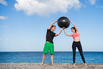 Sport und Fitness Instruktor zeigt junger Frau wie man mit Gymnastikball trainiert am Strand im Freien - obrazy, fototapety, plakaty