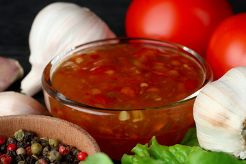 red sauce in a glass bowl near the red hot peppers and the leaves are green with vegetables on a black wooden background