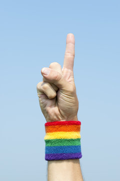 Hand of athlete with gay pride rainbow colors wristband pointing number one finger up into bright blue sky