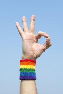 Hand of athlete giving OK sign with gay pride rainbow colors wristband against bright blue sky