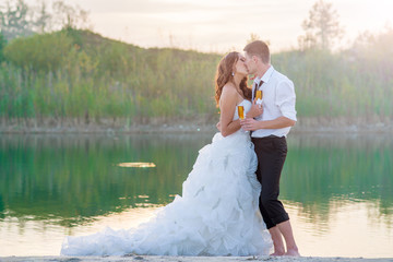 Romantic young and happy bride kissing on the lake with a glass of wine at sunset. Horizontal