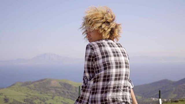 Waist Up Rear View of Woman with Curly Afro Hairstyle Wearing Black and White Plaid Shirt and Admiring View of Water and Island from Elevated Observation Point on Sunny Day.