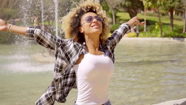 Happy young woman in sunglasses rejoicing with outspread arms as she relaxes in front of a fountain