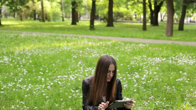A young woman is sitting on the grass in the park with a tablet and writing messages, touching the screen and smiling