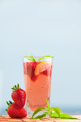 Glass of strawberry lemonade with pieces of strawberry, lemon and fresh mint. Closeup of mojito with beach on background. Fresh summer bright strawberry cocktail.