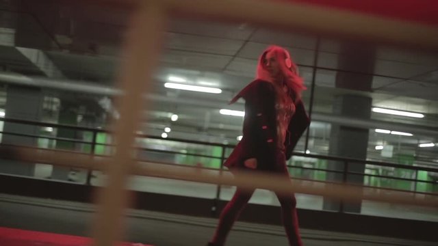 Teenage girl with long blonde hair walking fast along the street,listening to the music,wearing headphones at night under red lights, wearing black coat, jeans and high heel boots. 