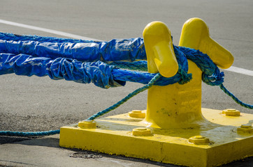 Heavy blue ropes of an ocean-going ship wrap around a yellow mooring bollard on a city pier in the harbor. - 111203499