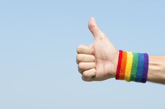 Hand of athlete giving thumbs up with gay pride rainbow colors wristband against bright blue sky