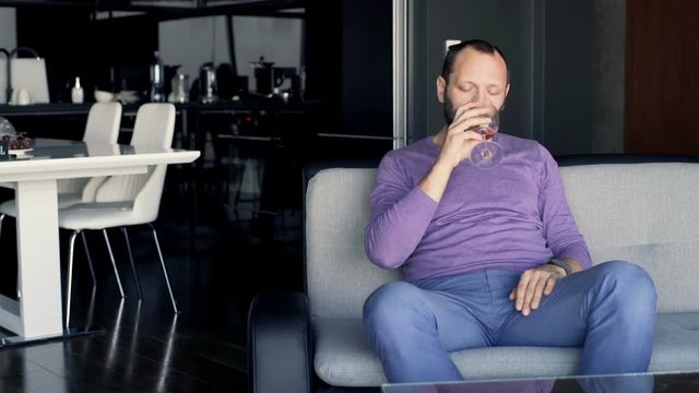 Sad, drunk man drinking red wine sitting on sofa at home
