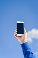 Holding mobile cell phone over blue sky white clouds background.