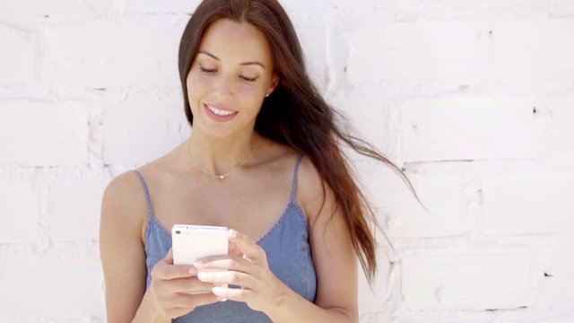 Pretty young woman leaning back against a white stone wall with her hair blowing in the breeze checking a text message on her smartphone