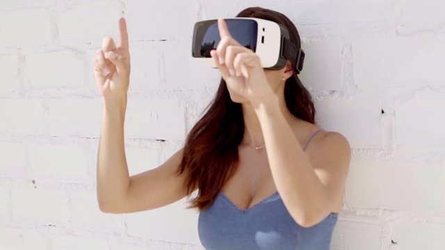 Young woman wearing virtual reality goggles leaning against a white brick wall in summer sunshine gesturing with her hands as she explores her virtual environment